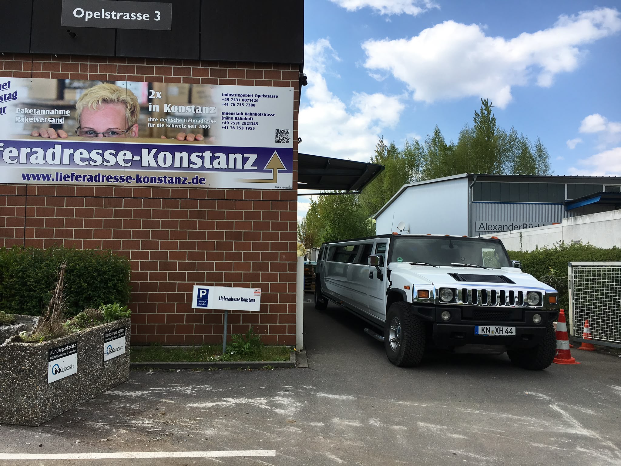 Hummer H2 Stretchlimousine bei der Lieferadresse Konstanz, Filiale Opelstraße 3 Weiße Hummer H2 Stretchlimousine vor der Einfahrt der Lieferadresse Konstanz in der Opelstraße 3 mit Firmenschild und Kundenparkplätzen